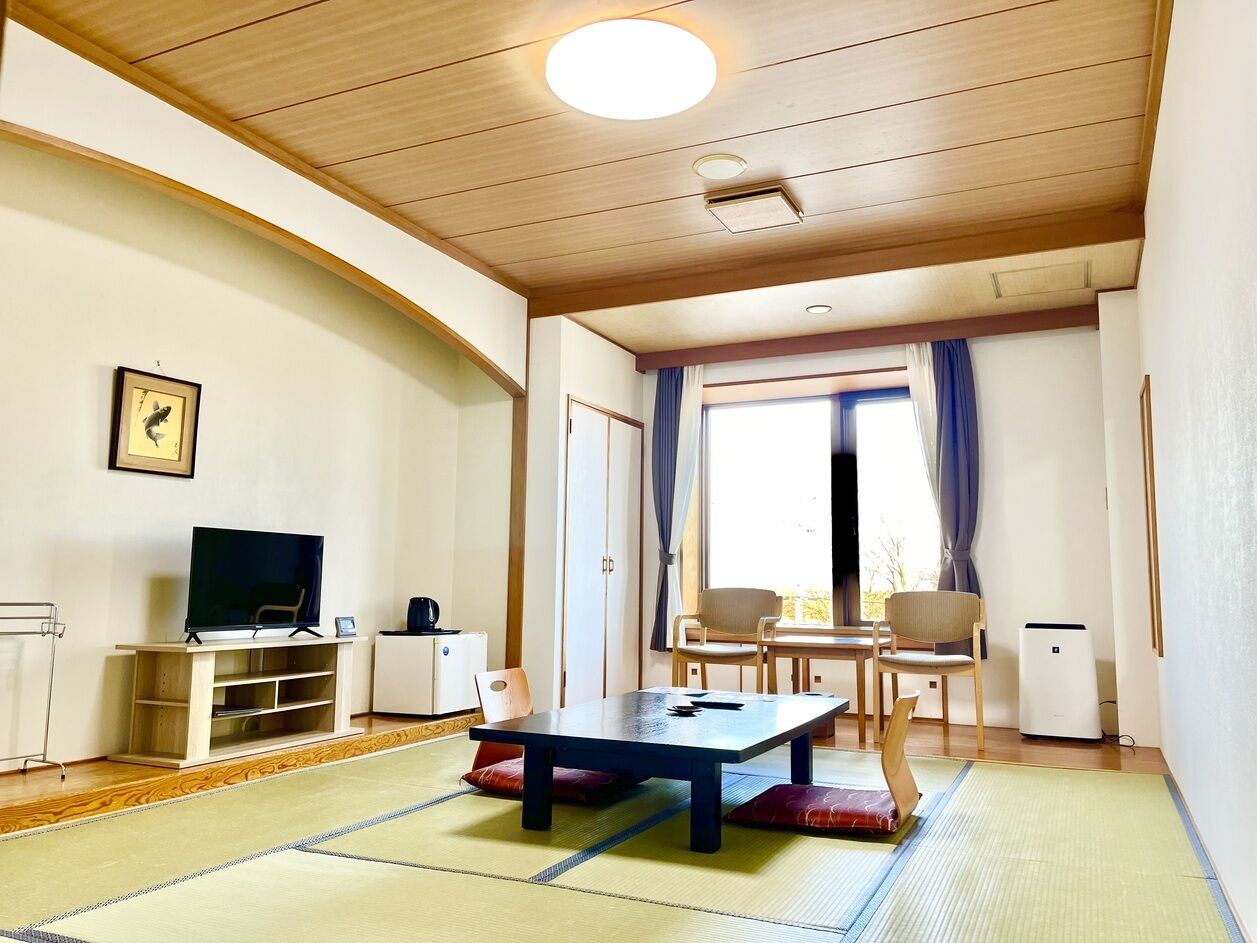 Ein Zimmer mit Tatami-Matten, einem Fernseher, einem Tisch, Stühlen und einem fensterbepflanzten Fenster.