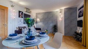 Ein moderner Essbereich mit einem Glastisch, gedeckt für vier Personen, umgeben von Stühlen. Im Hintergrund befindet sich eine Couch und ein kleiner Tisch mit einer Lampe.