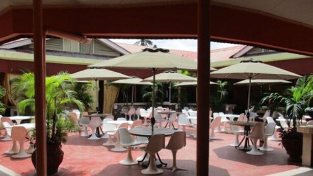 An outdoor dining area with multiple tables and chairs, covered by umbrellas, and surrounded by potted plants.