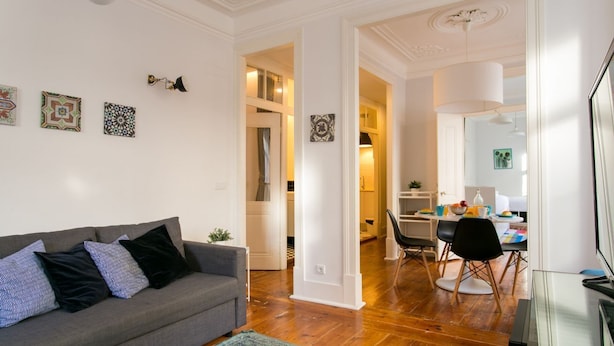 Een moderne woonkamer met een grijze bank, een eethoek met een tafel en stoelen, en een keuken op de achtergrond.