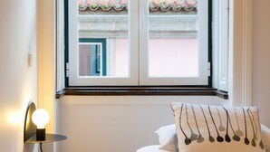 Een slaapkamer met een bed, een bedlampje en een raam met uitzicht op gebouwen.