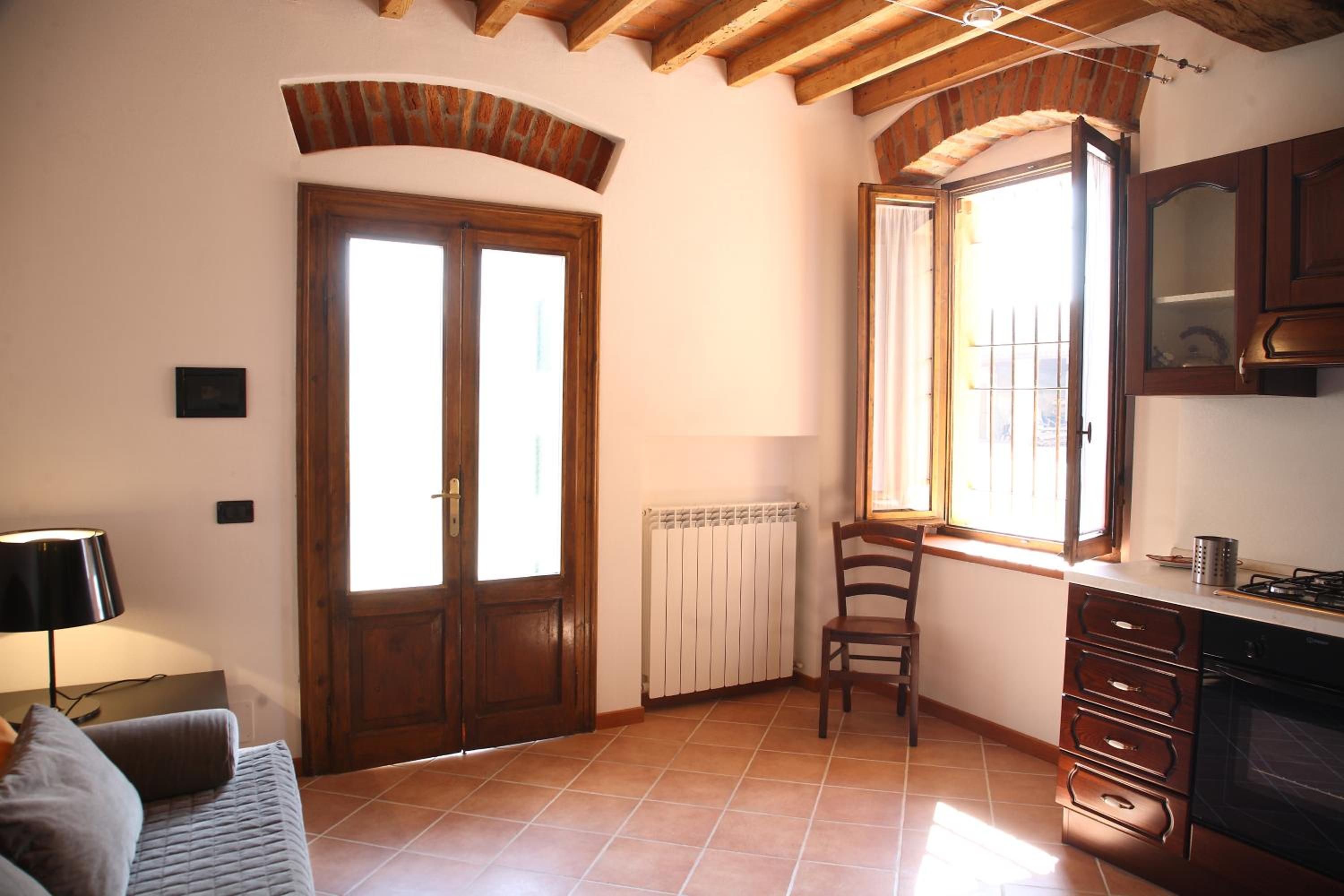 A room with a wooden door, a window with a view, a radiator, a chair, a sofa, and a kitchen area with cabinets and a stove.