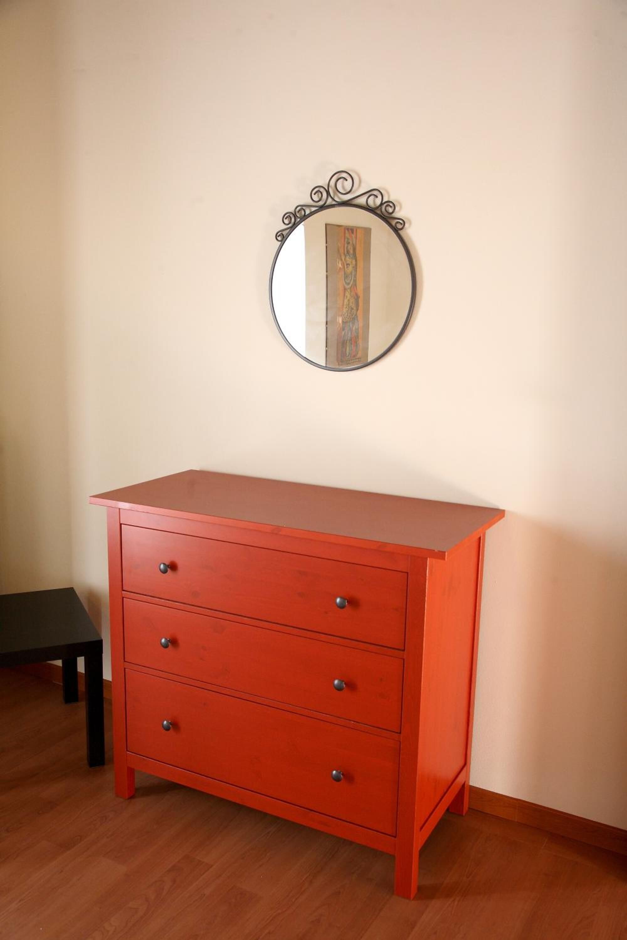 A red dresser with four drawers and a round mirror with a decorative frame.