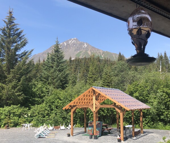 Un kiosque en bois doté d’un toit incliné, entouré d’une zone recouverte de gravier, avec des tables de pique-nique et des bancs.
