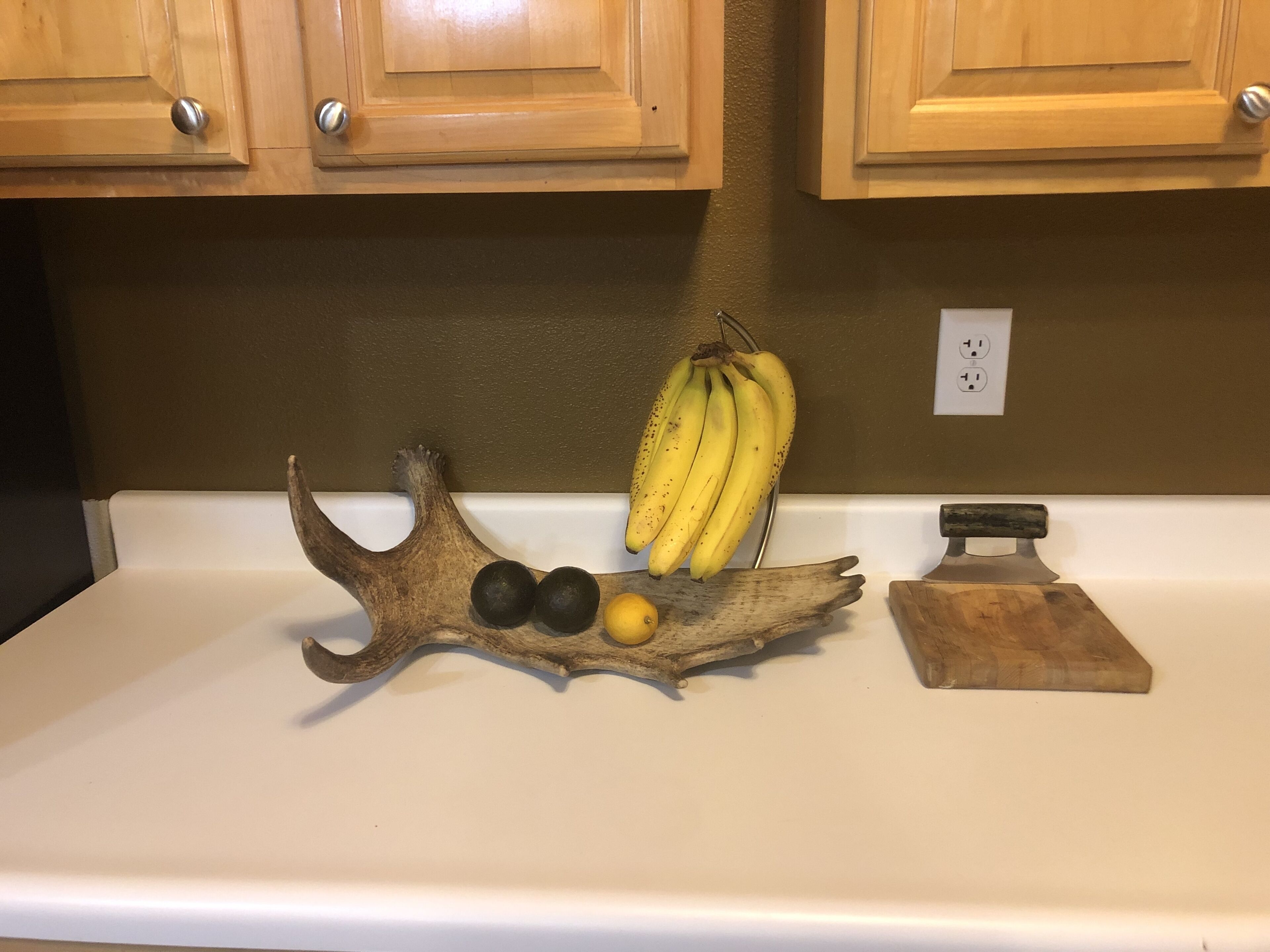 A kitchen counter with wooden cabinets, a white countertop, and a decorative piece with fruits and a knife.