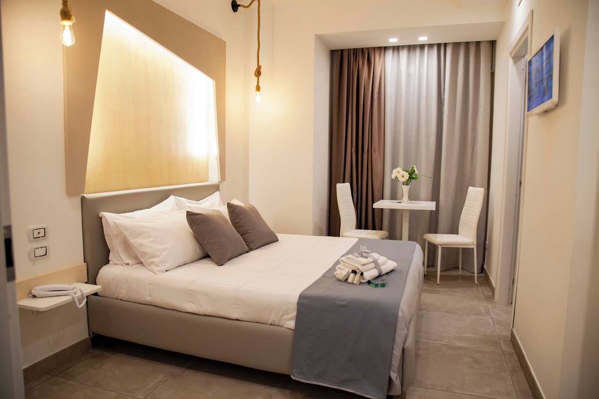 A neatly arranged hotel room with a large bed, a small table with a chair, and a wall-mounted lamp.