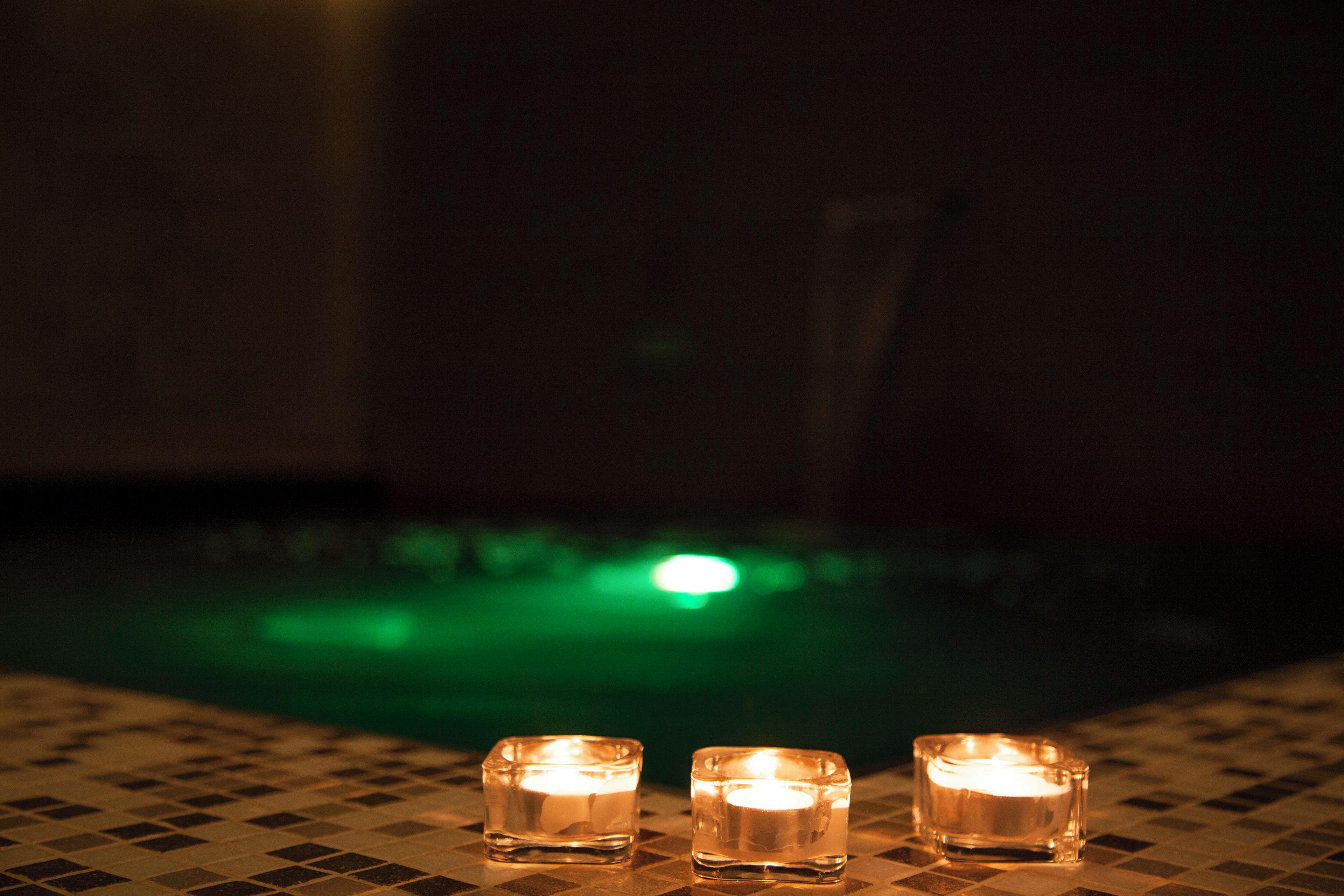 Drei brennende Kerzen auf einer gefliesten Oberfläche in der Nähe eines Pools, im Hintergrund ein grünes Licht.