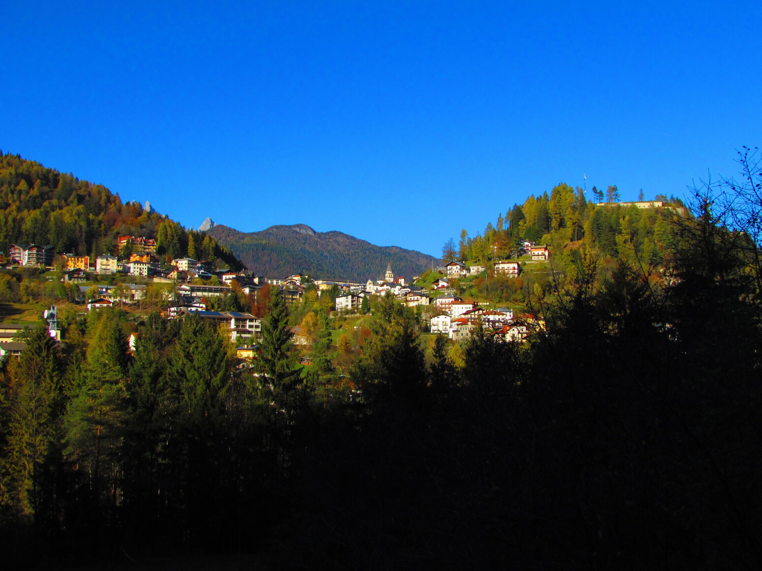 Un pueblo ubicado en un valle, con edificios rodeados de colinas boscosas y montañas al fondo.