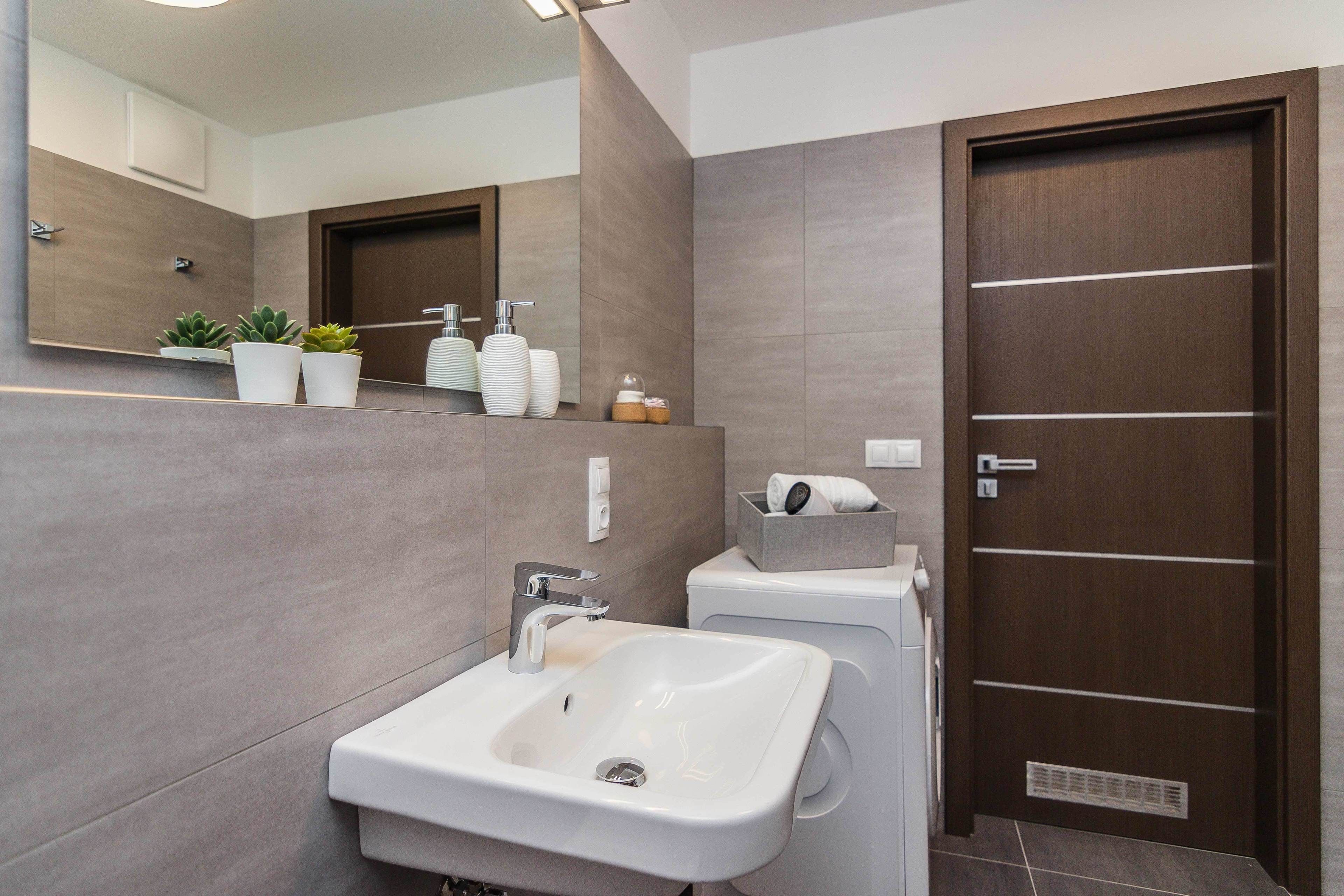 A modern bathroom with a white sink, a mirror, and a wooden door.