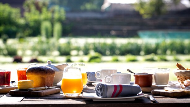 Une table de petit-déjeuner, dressée à l’extérieur, avec une variété d’aliments et de boissons, notamment un muffin, du jus d’orange et du lait, et offrant une vue sur la verdure et un bâtiment en arrière-plan.