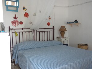 A bedroom with a double bed, a bedside table with a lamp, a small shelf with decorative items, and a wall-mounted fan.