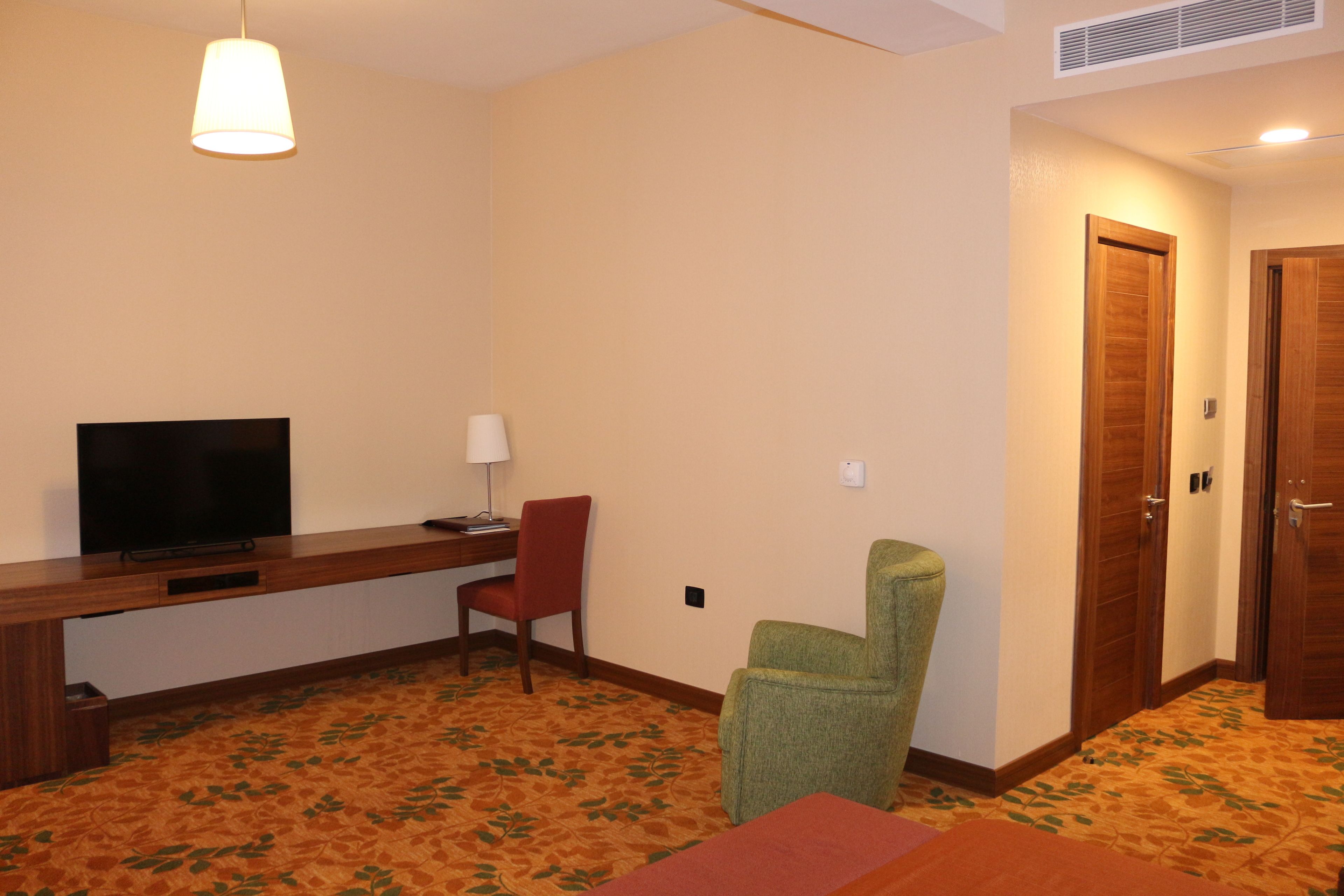 A hotel room with a wooden desk, a television, a red chair, a green chair, and a carpeted floor.