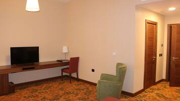 A hotel room with a wooden desk, a television, a red chair, a green chair, and a carpeted floor.