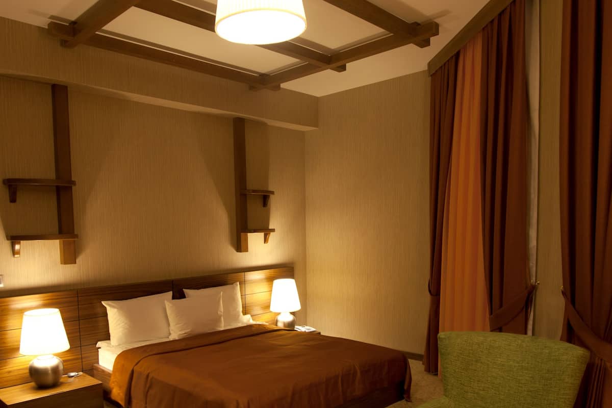 Habitación de hotel con cama, dos lámparas de noche, un sillón verde, una mesa de centro de madera y un ventanal con cortinas.