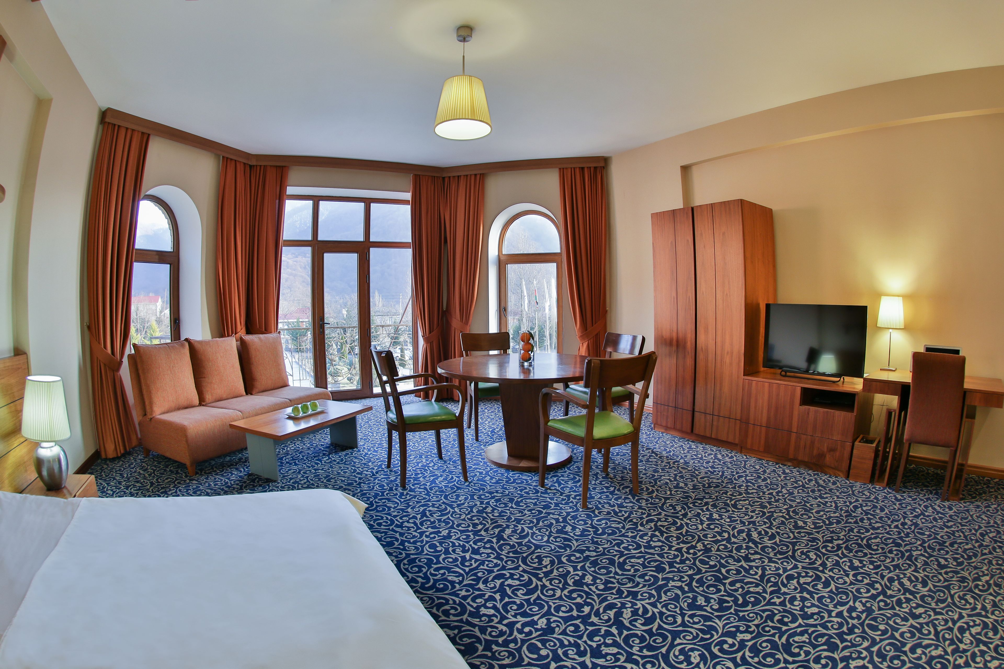 A hotel room with a bed, a sofa, a dining table with chairs, a TV, and large windows with curtains.