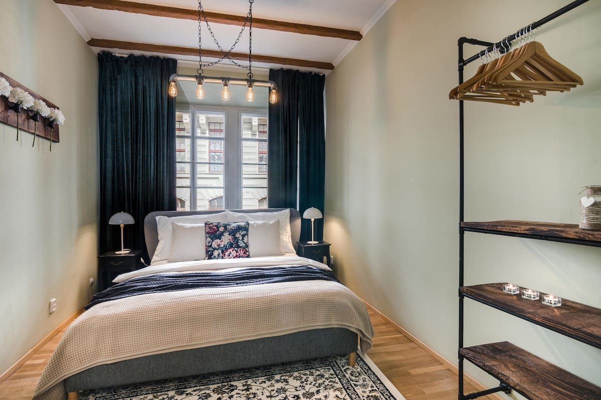 A bedroom with a bed, a wooden floor, a window with curtains, a hanging light fixture, and a metal shelf with candles.