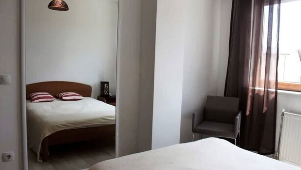 A bedroom with a bed, a chair, a nightstand, and a window with curtains. - Emerald Apartment (Pristina)