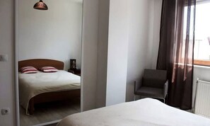 A bedroom with a bed, a chair, a nightstand, and a window with curtains. - Emerald Apartment (Pristina)