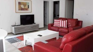 A modern living room with a red leather sofa, a white coffee table, and a flat-screen TV on a grey entertainment unit. - Emerald Apartment (Pristina)