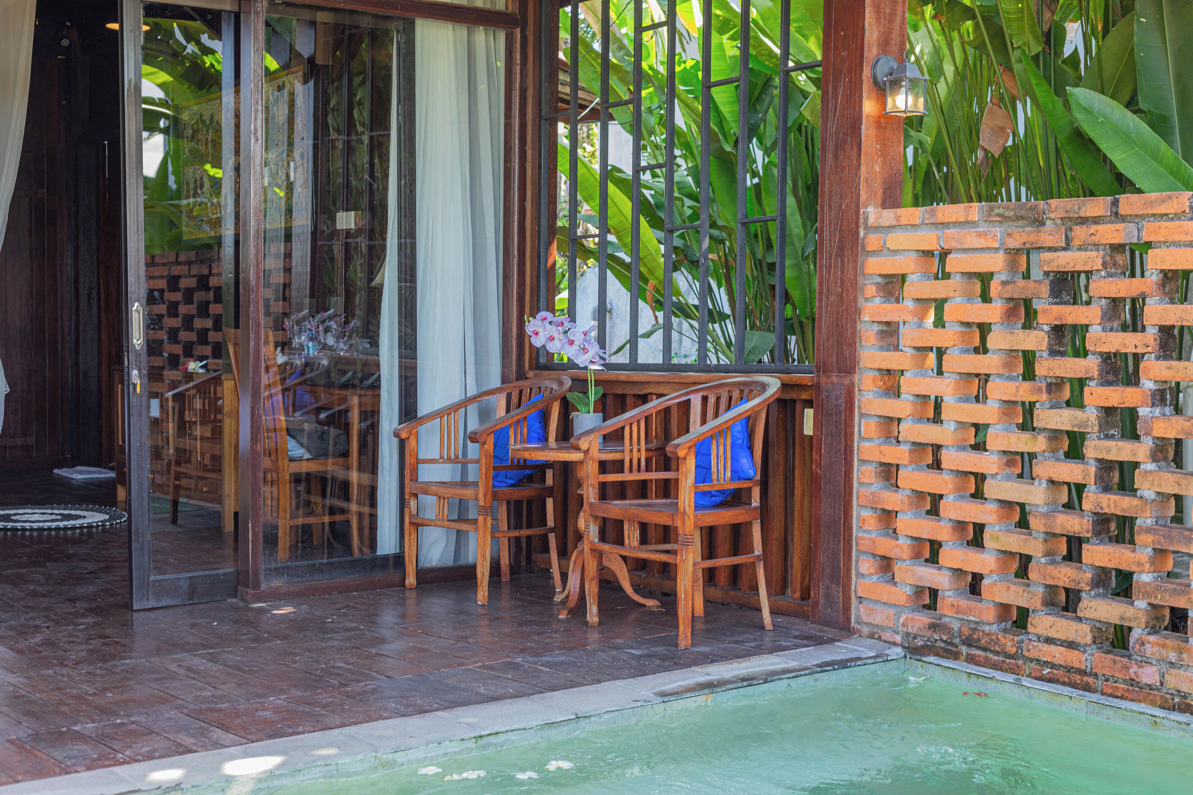 A wooden outdoor seating area with chairs and a table, a brick wall, and a glass door leading inside.