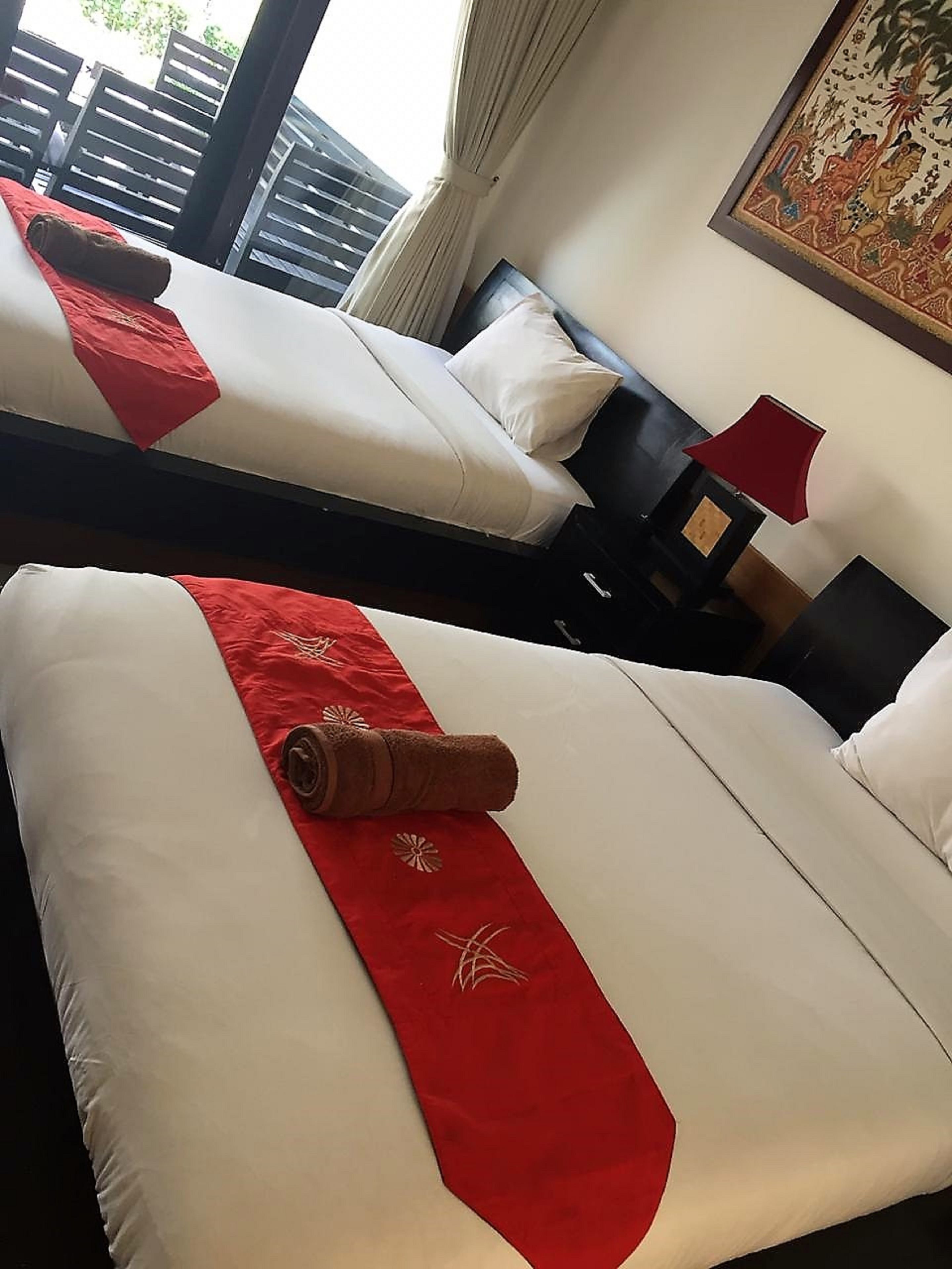 A hotel room with two beds, each with white linens and a red runner. There is a red lamp on a bedside table and a framed picture on the wall.