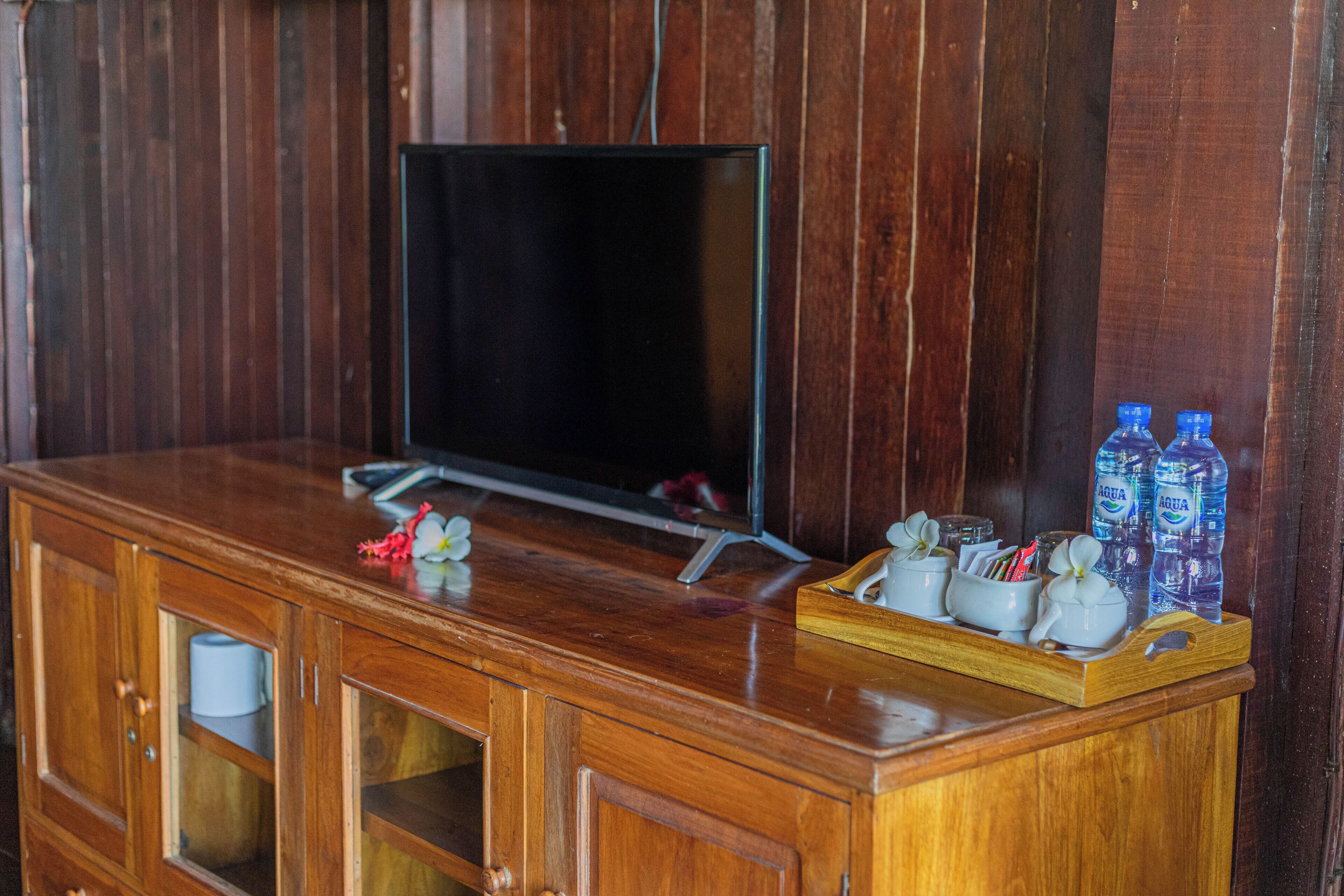 Een houten tv-meubel met een televisie, een dienblad met kleine witte bekertjes en een bloem, plus twee waterflessen.