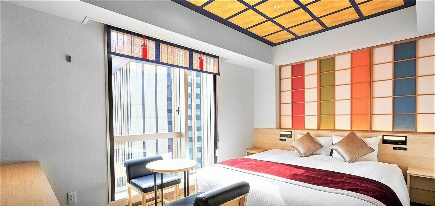 Habitación de hotel con cama, una mesita, una silla, un ventanal grande y un panel de pared de estilo tradicional japonés.