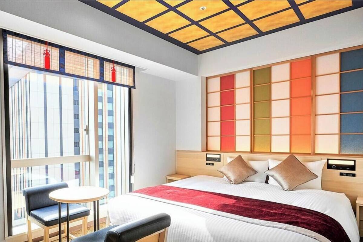Habitación de hotel con cama, una mesita, una silla, un ventanal grande y un panel de pared de estilo tradicional japonés.