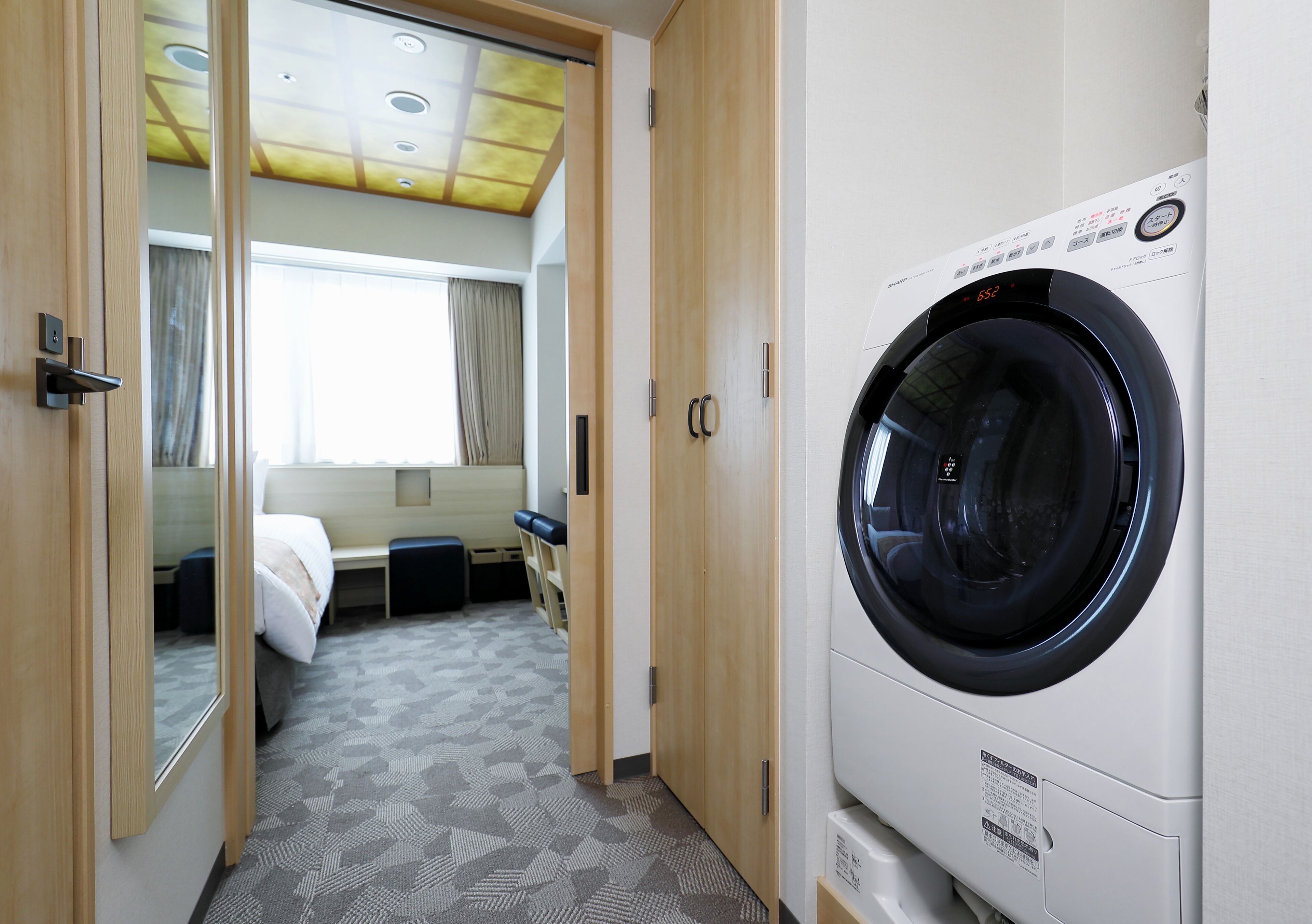 A hotel room with a washing machine, a bed, a bench, and a window with curtains.