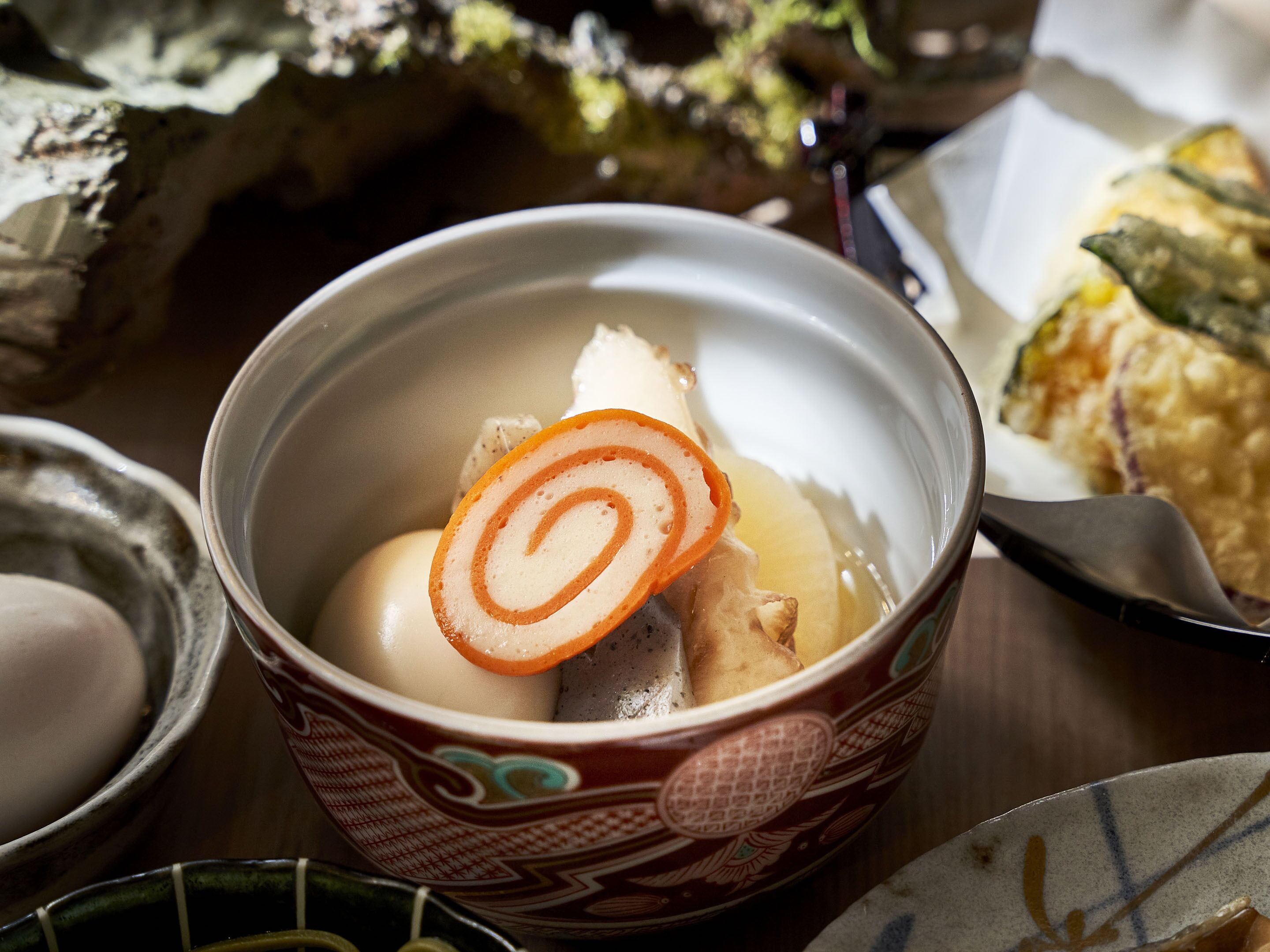 A bowl of soup with a slice of carrot and a piece of meat, surrounded by other dishes.