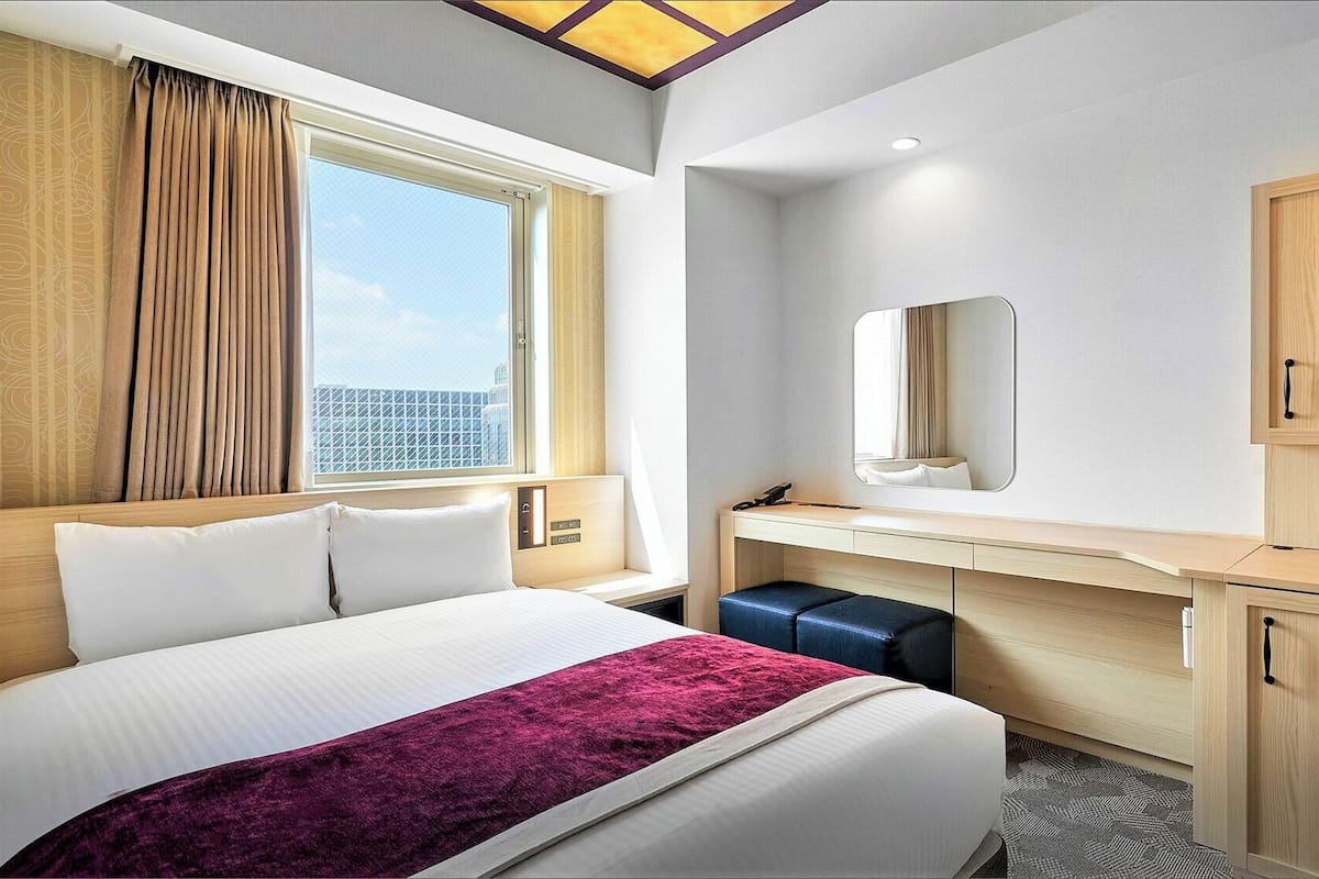 Una habitación de hotel con cama, escritorio, espejo y ventana con cortinas.