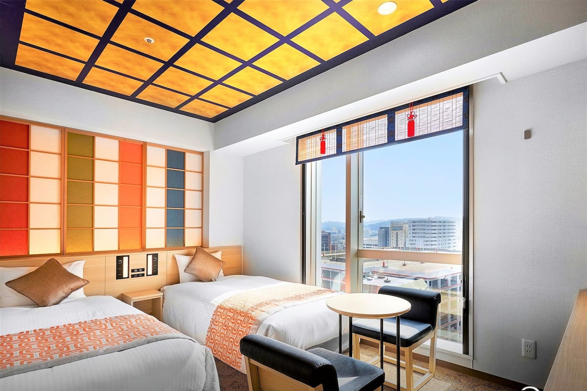 Habitación de hotel con dos camas, una mesita redonda y vistas de la ciudad a través de amplias ventanas.