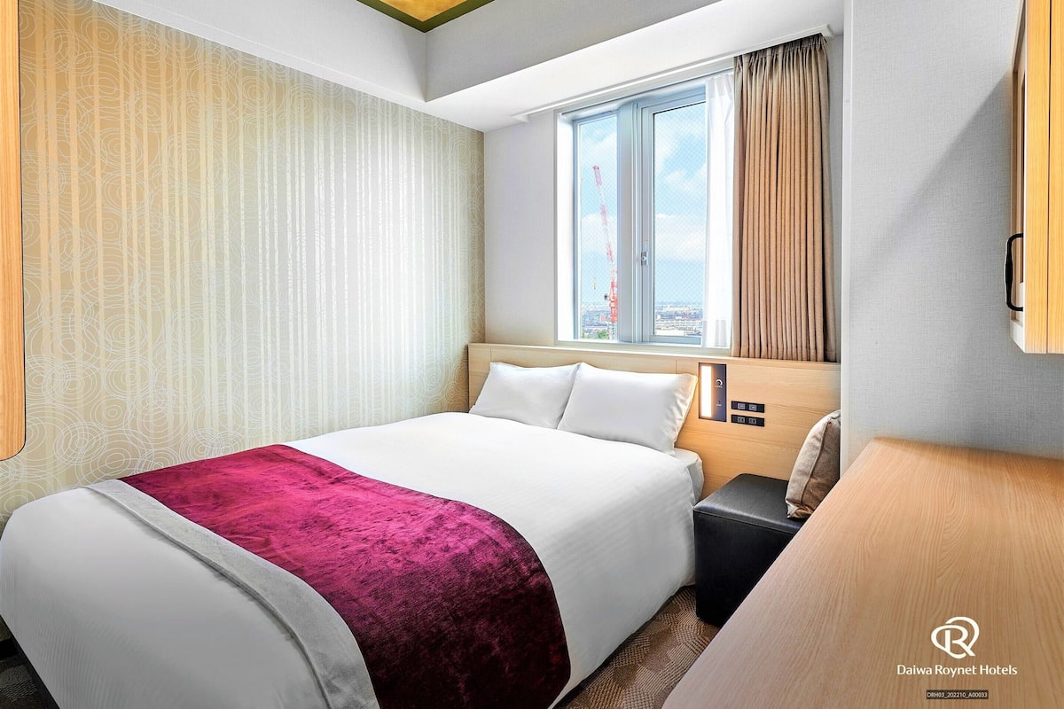 Habitación de hotel con cama, mesita de noche, cabecero de madera, ventana con cortinas y vistas a la ciudad.