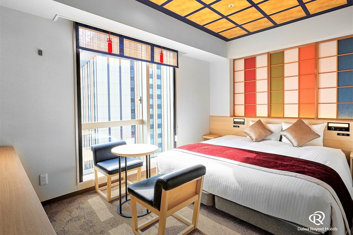 Habitación de hotel con cama, mesitas de noche, una pequeña mesa redonda con sillas, una ventana con persianas y un techo con diseño de paneles de madera.