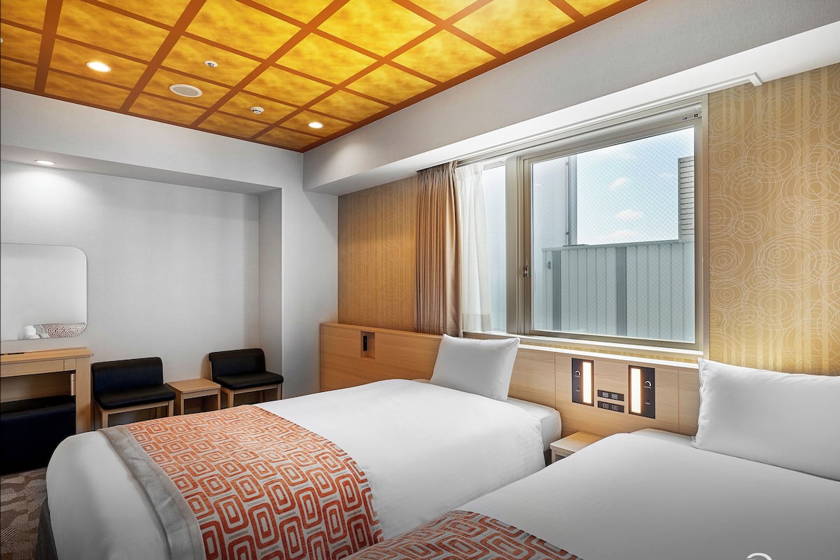 A hotel room with a large bed, a desk, two chairs, a window with curtains, and a ceiling with recessed lighting.