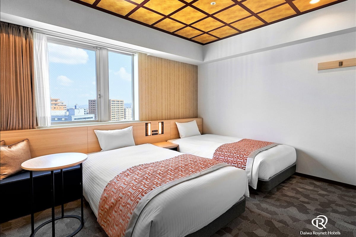 Habitación de hotel con dos camas, una mesita, un sofá y ventana con vista a la ciudad.