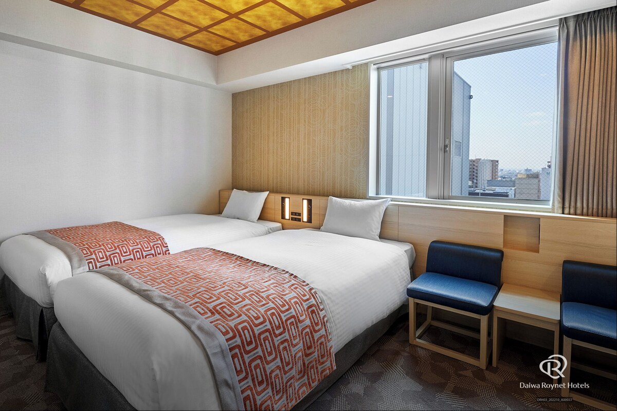 Une chambre d’hôtel avec deux lits, une fenêtre donnant sur la ville et un coin salon avec des fauteuils.