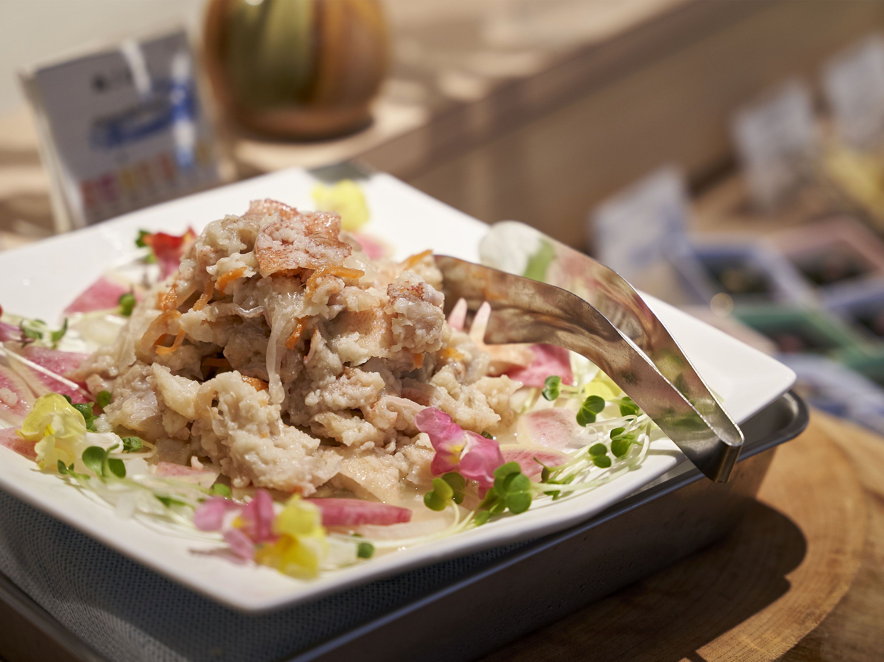 A square plate with a dish of food, a fork, and some flowers.