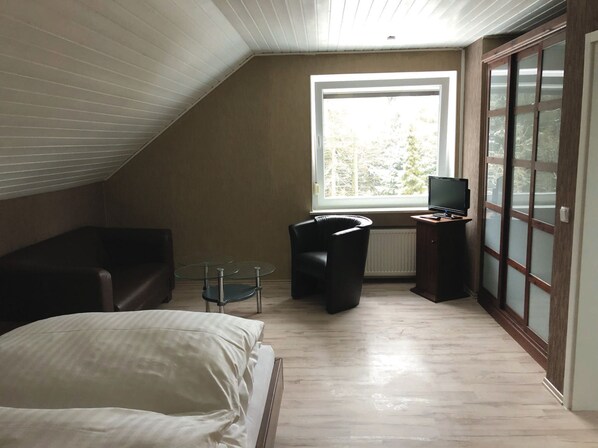 A bedroom with a bed, a chair, a small table, a TV, and a window with a view of trees.