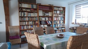 A dining area with a table covered by a tablecloth, surrounded by wicker chairs. Behind the table, there are two large wooden bookshelves filled with books.