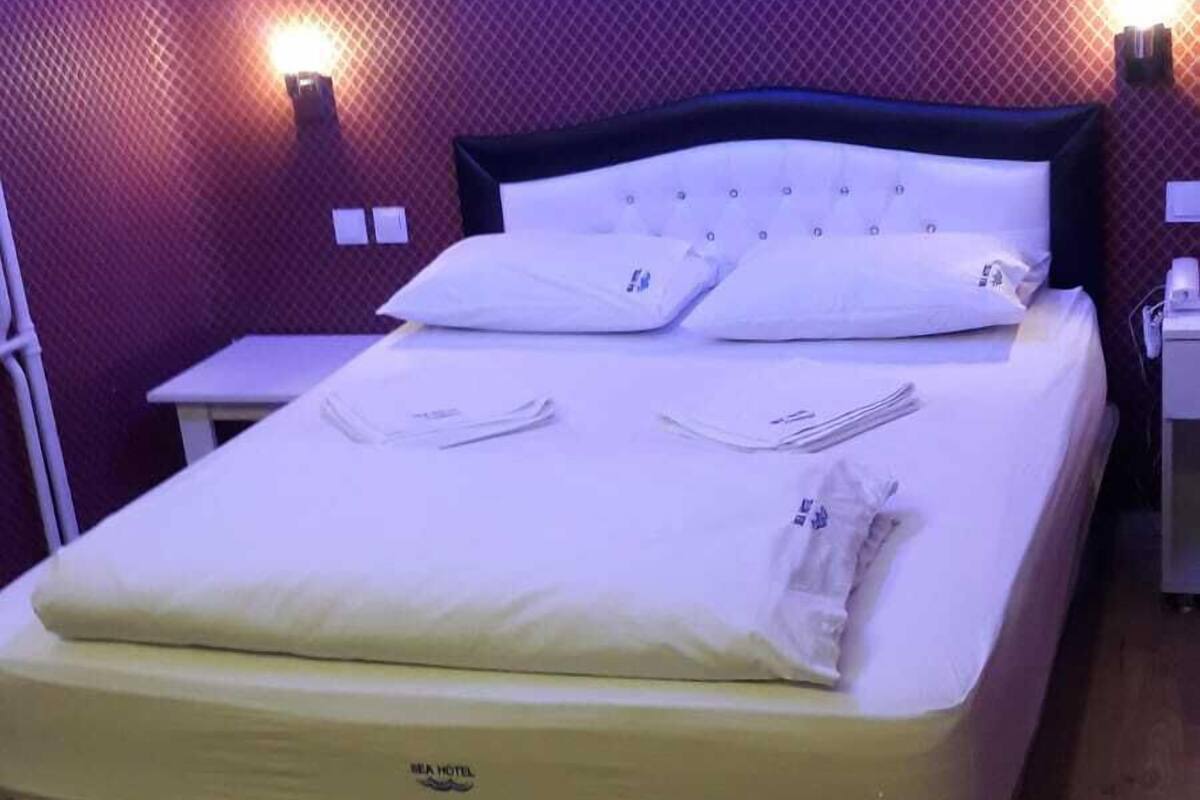 A neatly made bed with a mattress labeled "WILHA HOTEL" in a room with purple walls and two bedside lamps.