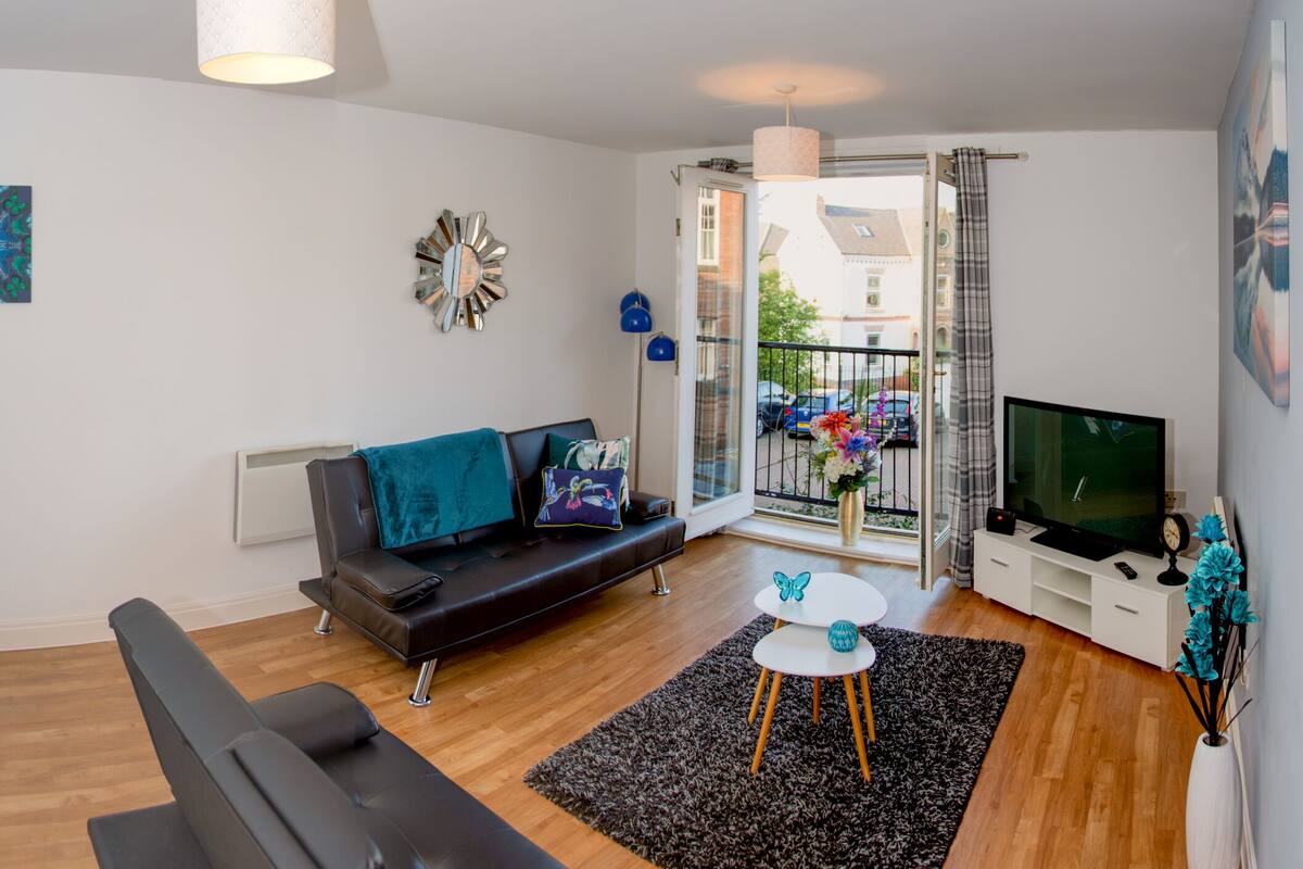 A modern living room with a black leather sofa, a small white table, a TV, and a balcony with a view of parked cars and buildings.