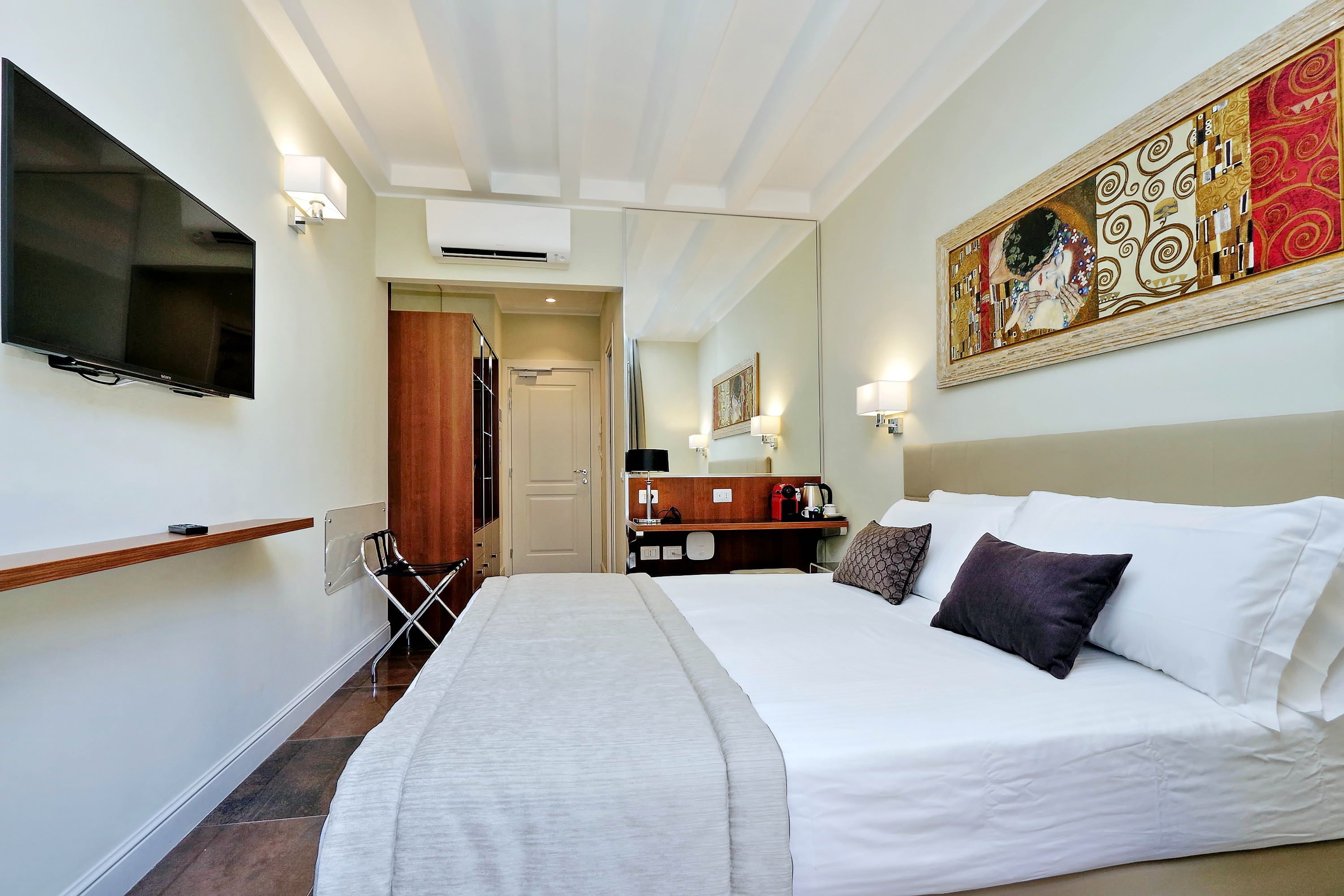Habitación de hotel con una cama grande, un televisor de pantalla plana montado en la pared, un cuadro sobre la cama y un escritorio de madera con una silla.
