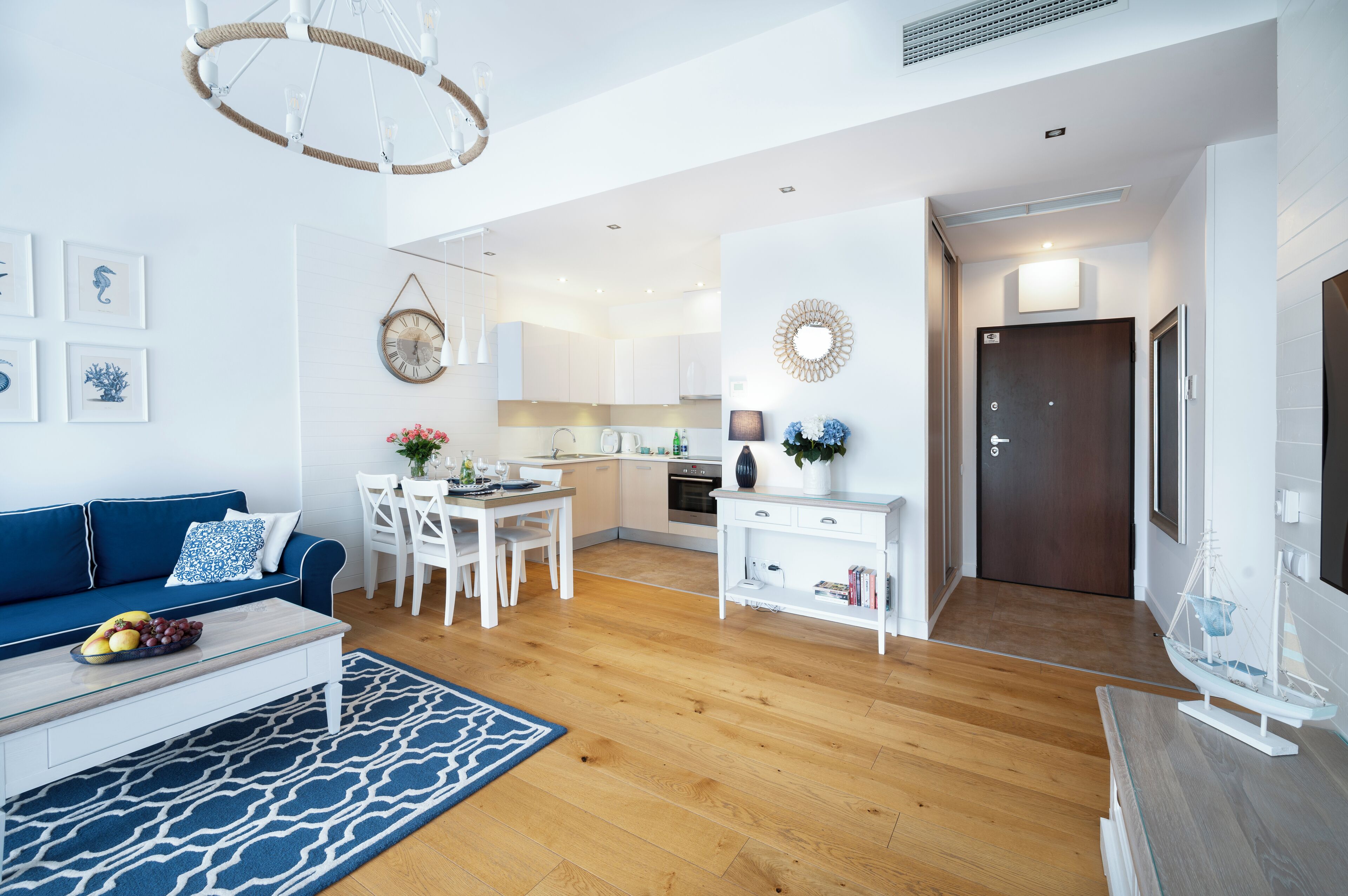 A modern living room with a blue sofa, white coffee table, and a dining area with a white table and chairs. A kitchen is visible in the background with white cabinets and a wooden countertop.
