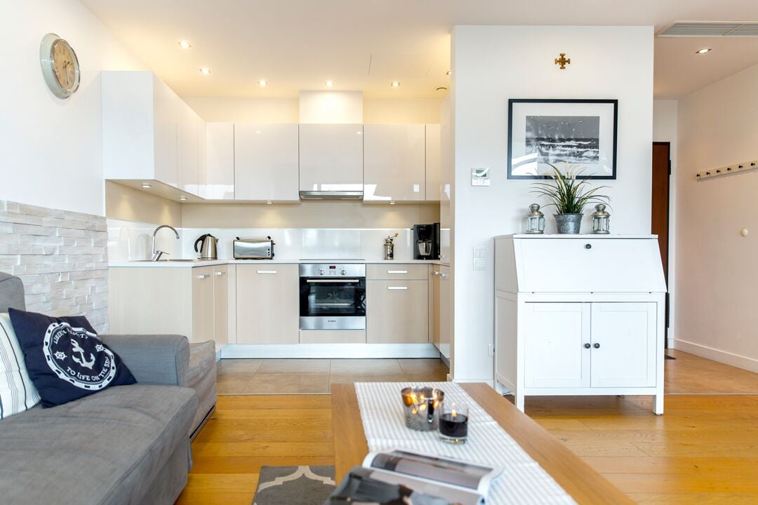 Una cocina moderna con armarios blancos, horno empotrado y cafetera. Cuenta con un comedor con mesa de madera y sofá.