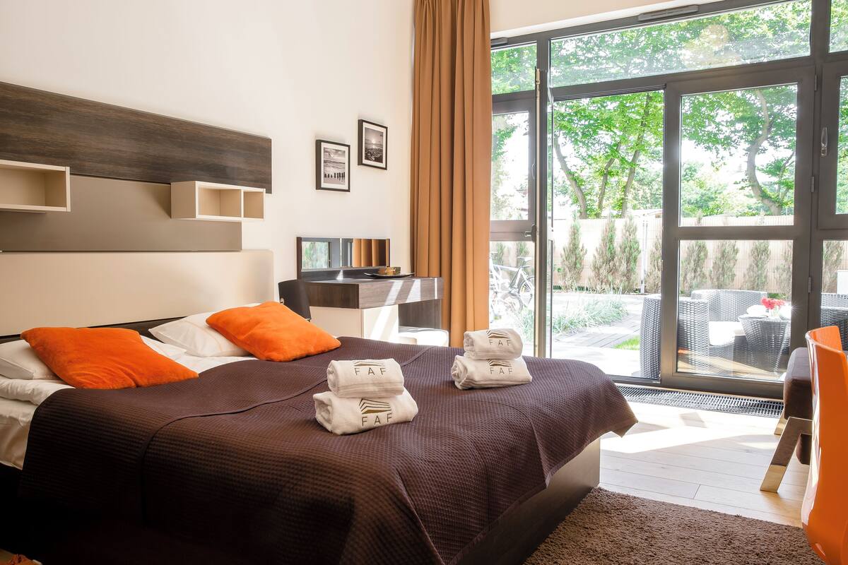 Habitación de hotel moderna con una cama grande, almohadas anaranjadas, un escritorio con una computadora y vistas al exterior a través de amplios ventanales.