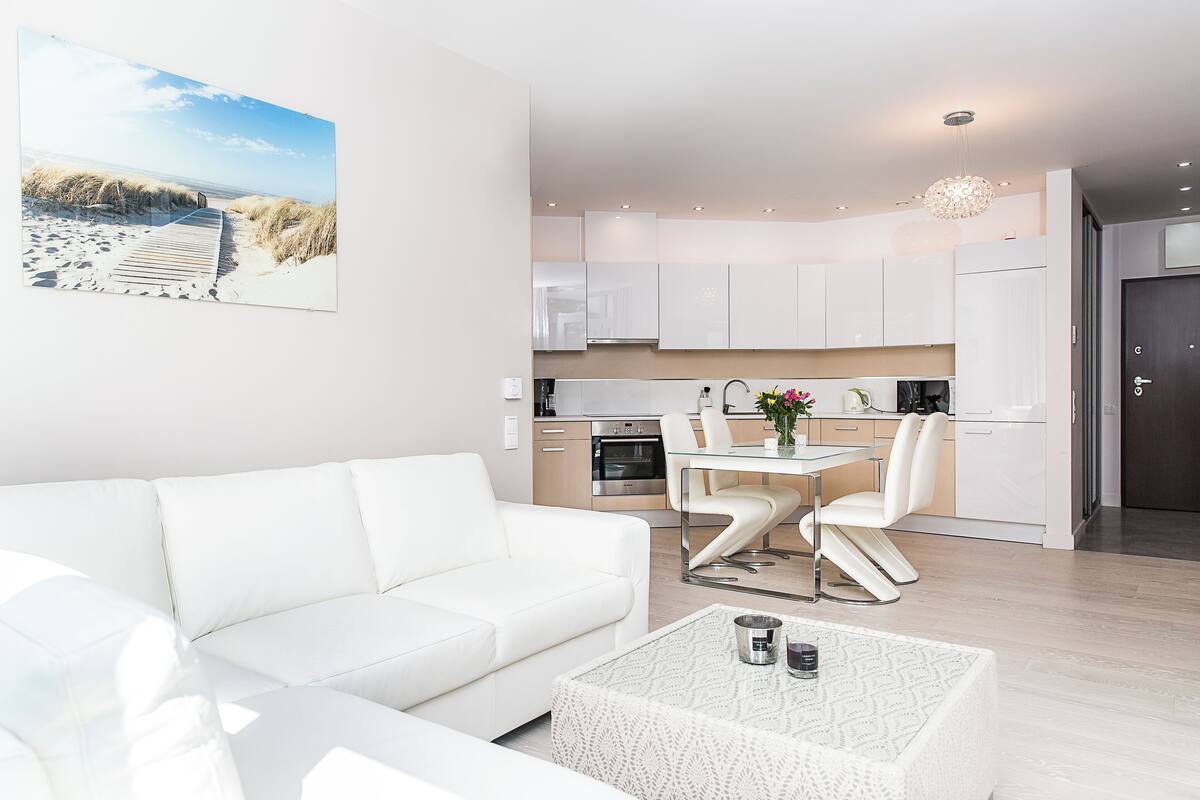 Una sala de estar moderna con un sofá blanco, una mesa de comedor con sillas y una cocina con armarios y electrodomésticos blancos.