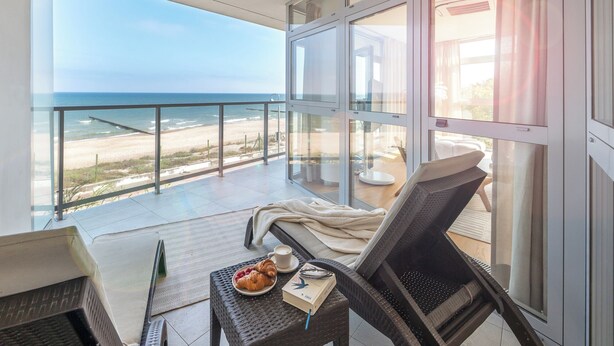 Un balcon avec vue sur la plage et l’océan. On y trouve une chaise en osier, une petite table avec un croissant, une tasse et un livre.
