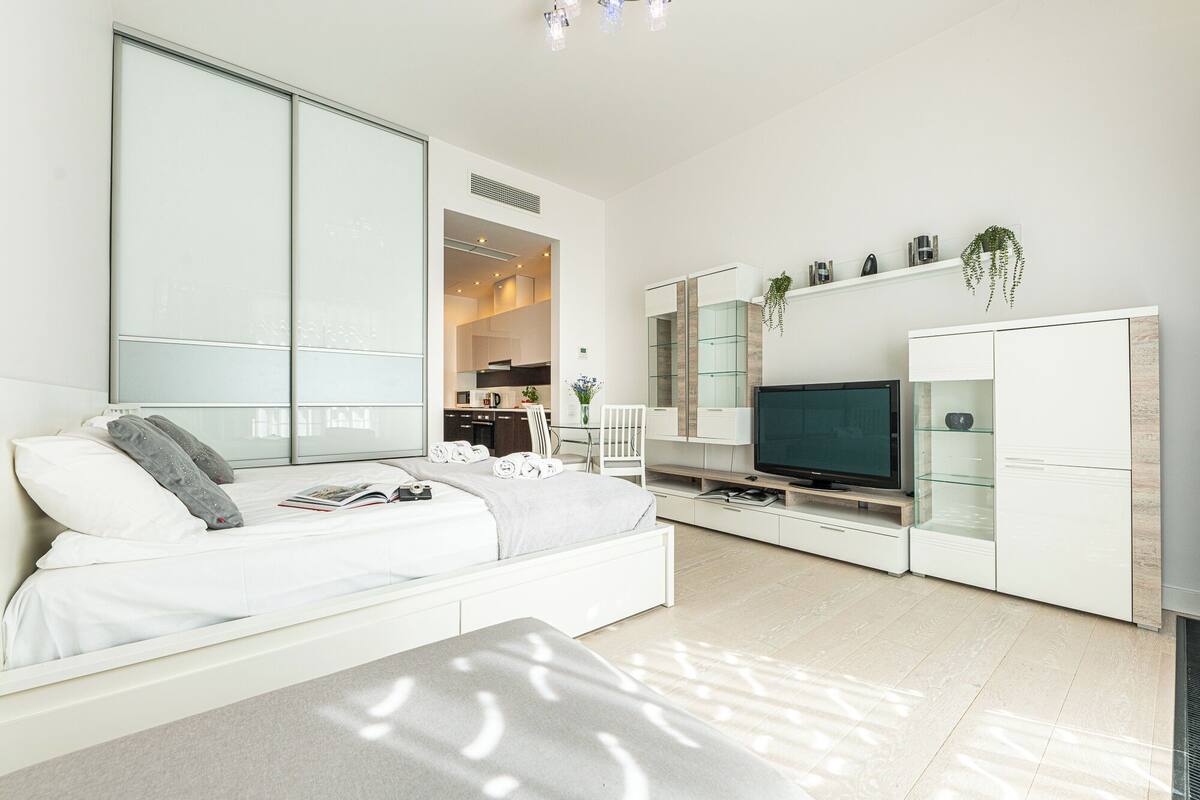 Una sala de estar moderna con un sofá blanco, un televisor de pantalla plana y una cocina visible a través de un panel de vidrio.
