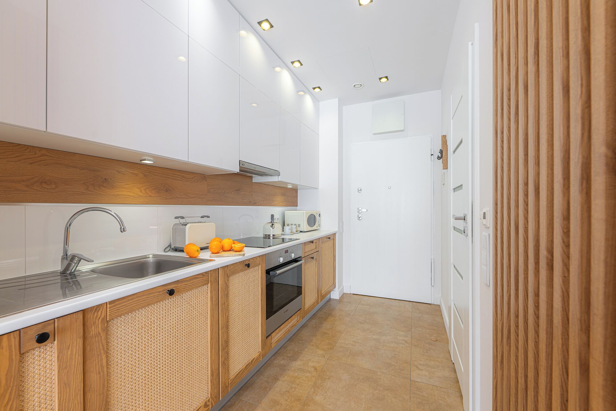 A modern kitchen with white cabinets, a stainless steel sink, a microwave, and a toaster. There are oranges on the counter and a wooden panel on the wall.